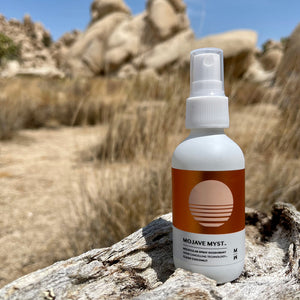 
                  
                    A spray bottle of Mojave Myst Molecular Spray Deodorant resting on an old Joshua Tree trunk. Desert grasses and landscape are in the background.
                  
                